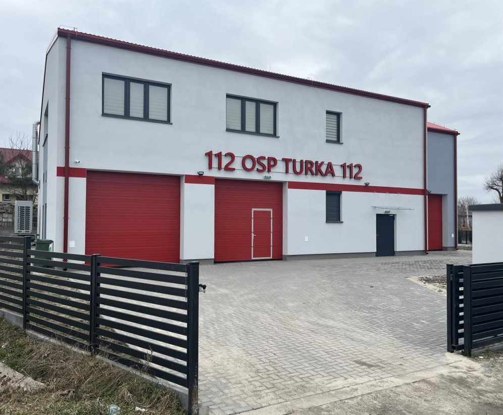 Budynek straży pożarnej z dużymi, czerwonymi bramami garażowymi i napisem "112 OSP TURKA 112" na elewacji. Obiekt jest dwukondygnacyjny, z białymi ścianami i płaskim dachem.
