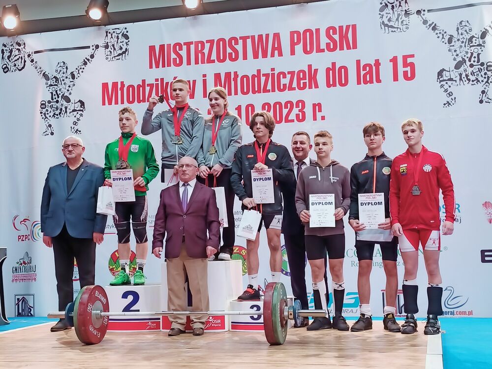 Grupa młodzieży i dorosłych stoi na podium i obok niego na tle baneru z napisem "Mistrzostwa Polski Młodzików i Młodziczek do lat 15". Na scenie znajduje się również sztanga.