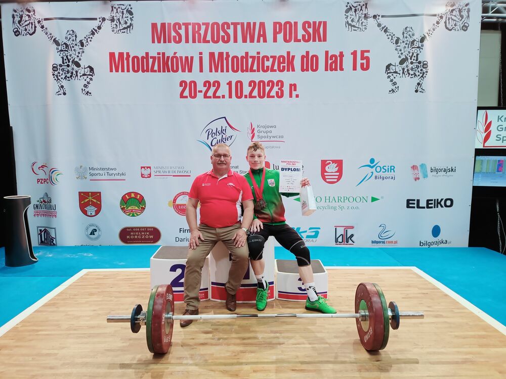 Na zdjęciu dwie osoby stoją po obu stronach sztangi na tle plakatu z napisem o mistrzostwach Polski w podnoszeniu ciężarów.