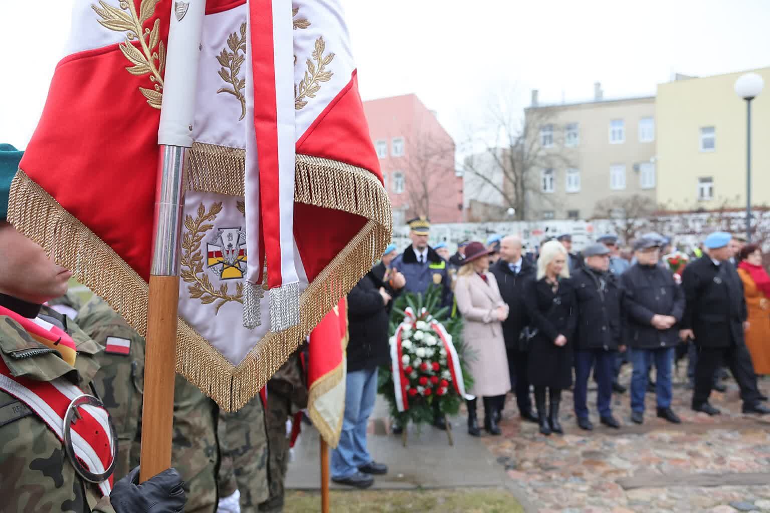 Osoba trzymająca sztandar z polskimi symbolami narodowymi; grupa ludzi w tle podczas oficjalnej uroczystości.