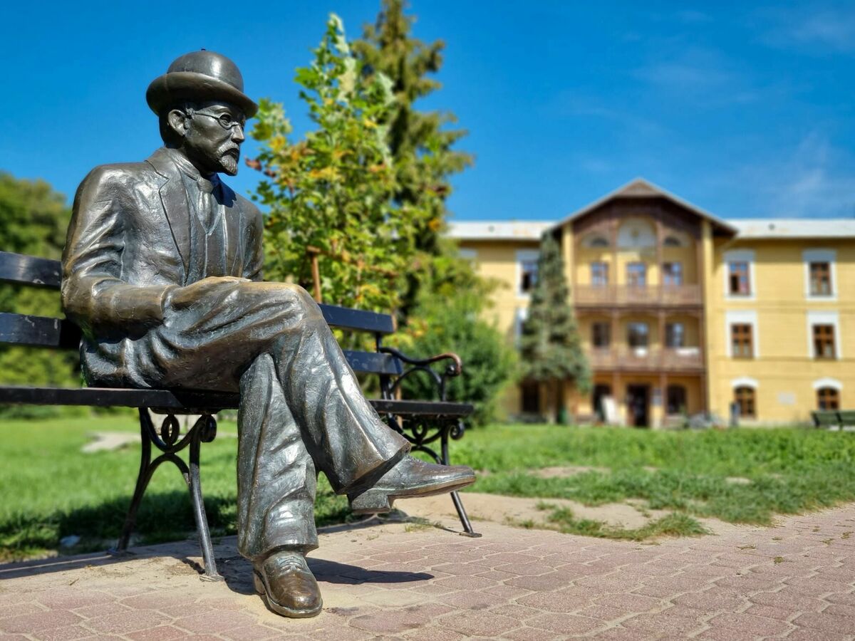 Pomnik mężczyzny siedzącego na ławce w parku, w tle zielone drzewa i żółty budynek.