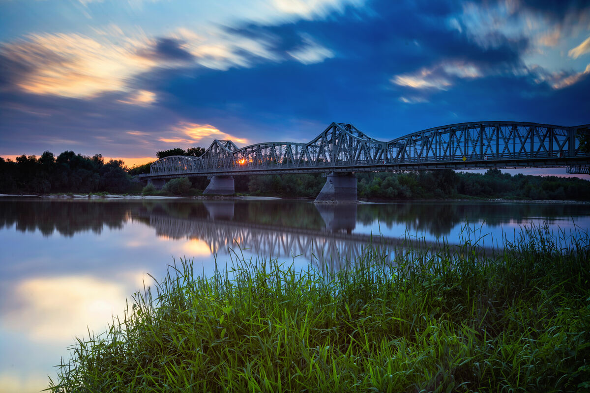 Zdjęcie przedstawia długi, metalowy most kolejowy nad spokojną rzeką odbijającą kolorowe niebo o zachodzie słońca, z zieloną trawą na pierwszym planie.