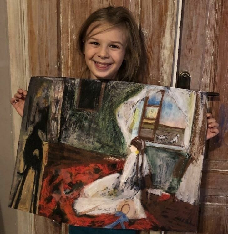 Dziewczynka trzyma kolorowy obraz przedstawiający abstrakcyjną scenę z postaciami. Uśmiecha się, stojąc przed drewnianymi drzwiami.